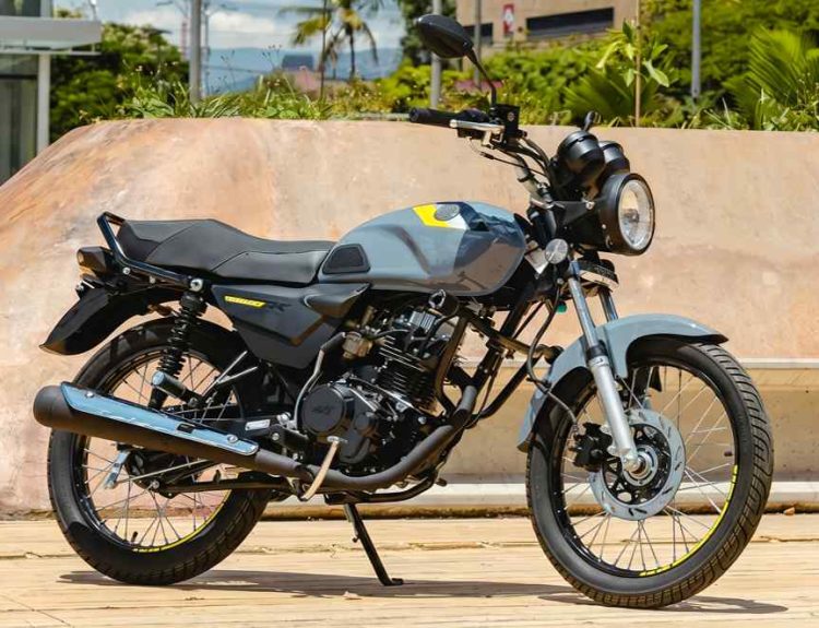 Motocicleta AKT AK125 NKD color gris nardo con detalles amarillos estacionada de perfil sobre una acera urbana, mostrando su diseño clásico con faro redondo y rines de radios.