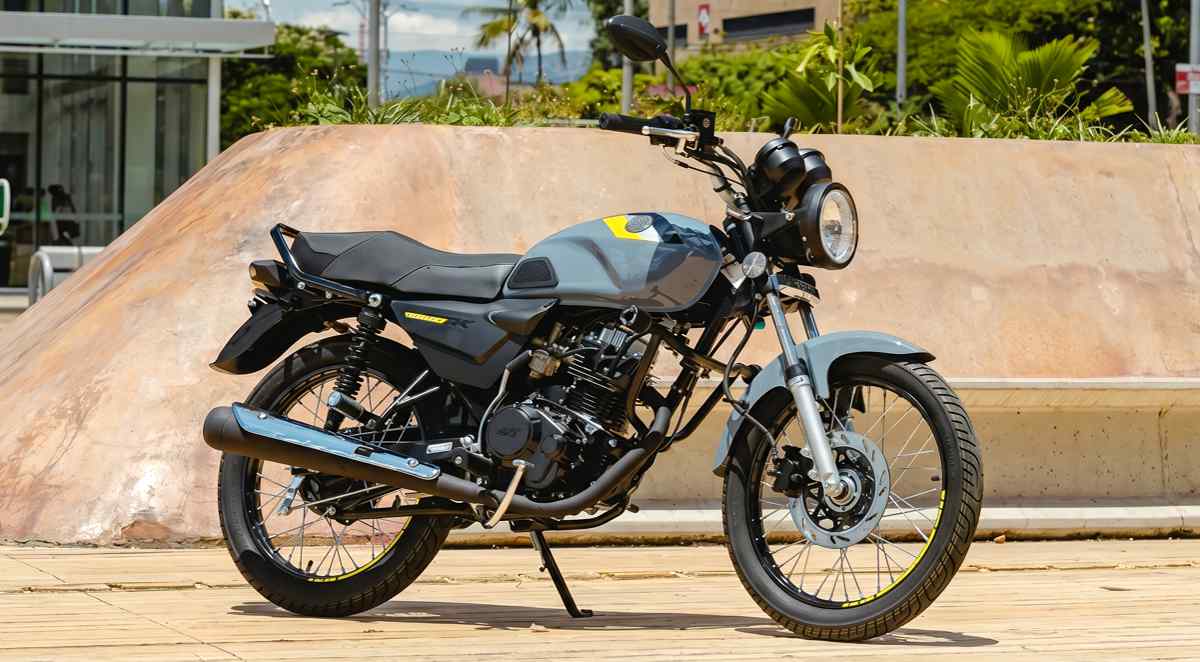 Motocicleta AKT AK125 NKD color gris nardo con detalles amarillos estacionada de perfil sobre una acera urbana, mostrando su diseño clásico con faro redondo y rines de radios.