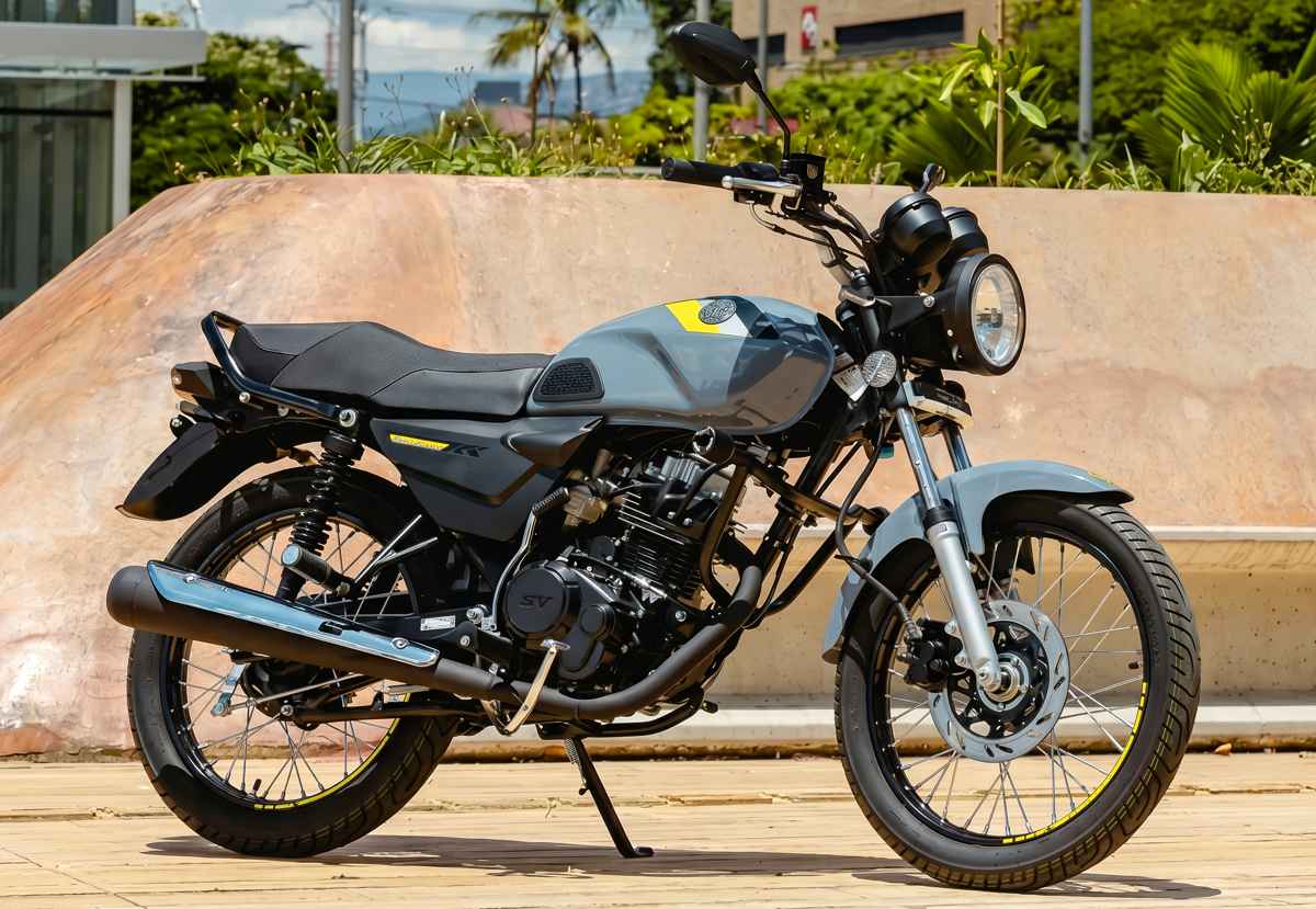 Motocicleta AKT AK125 NKD color gris nardo con detalles amarillos estacionada de perfil sobre una acera urbana, mostrando su diseño clásico con faro redondo y rines de radios.