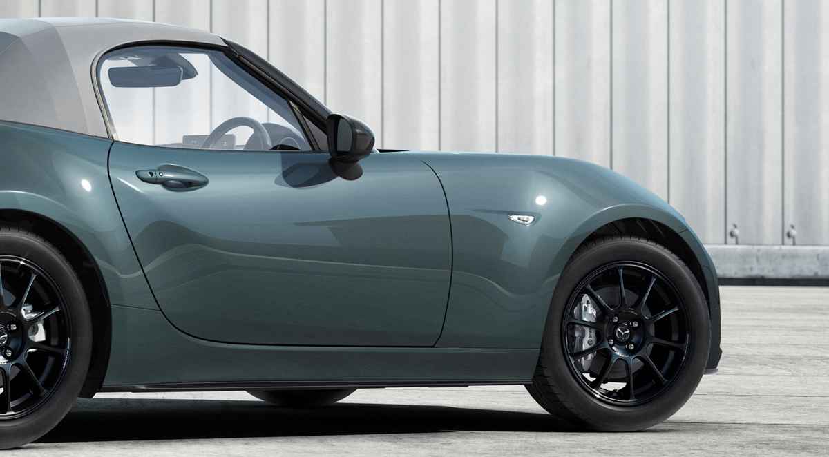 Vista lateral delantera del deportivo Mazda MX-5 convertible luciendo la nueva pintura exterior Verde Zinc, contrastando con sus rines negros y techo de lona gris.