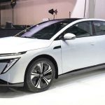 Vista frontal de tres cuartos del nuevo sedán eléctrico Nissan Primera EV en color blanco, exhibido en un salón del automóvil destacando su silueta aerodinámica y rines bitono.