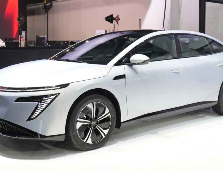 Vista frontal de tres cuartos del nuevo sedán eléctrico Nissan Primera EV en color blanco, exhibido en un salón del automóvil destacando su silueta aerodinámica y rines bitono.