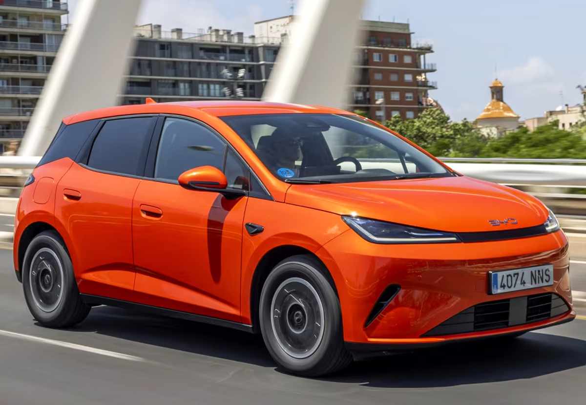 Vista frontal en movimiento del BYD Dolphin G DM-i híbrido enchufable color naranja recorriendo una vía urbana, mostrando su diseño aerodinámico.