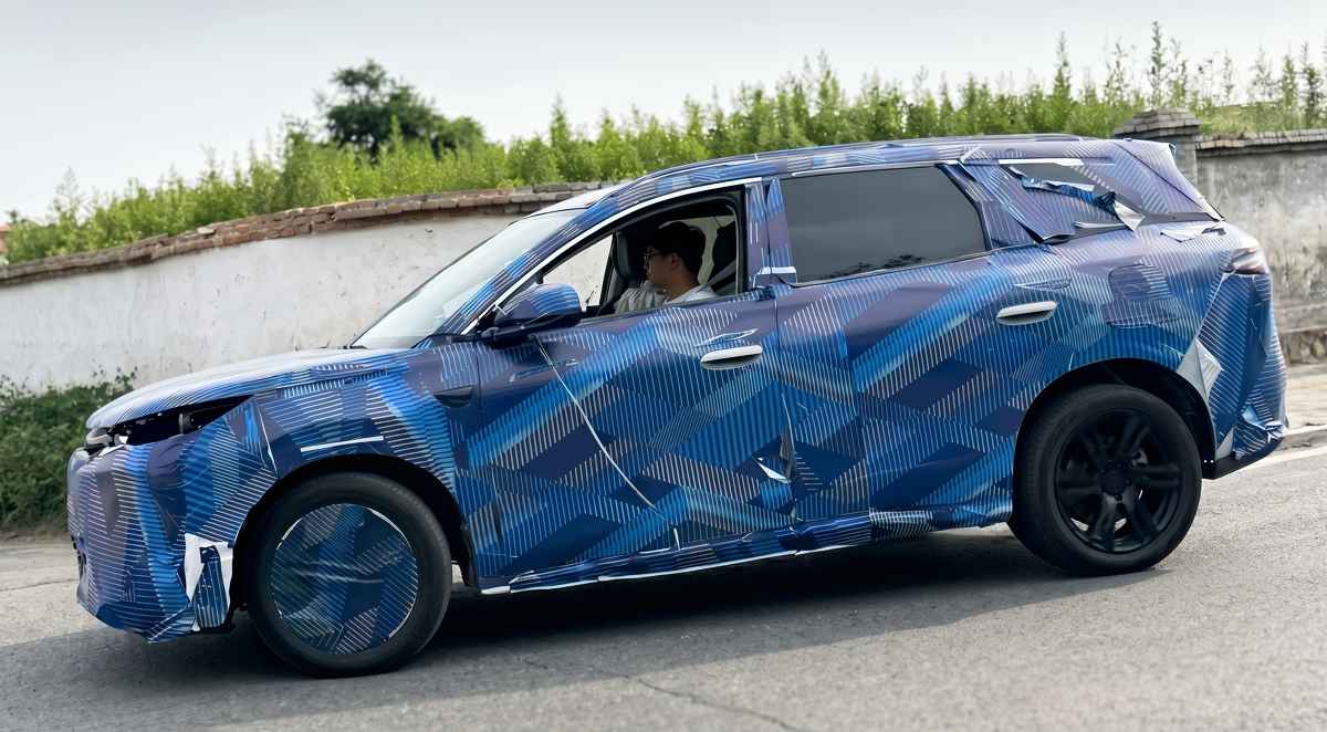 Vista de perfil en marcha de la nueva generación del BYD Tang cubierta con un denso camuflaje azul con patrones geométricos durante pruebas de ruta en China.