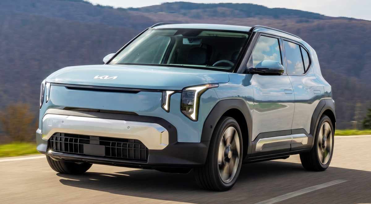 Vista frontal de tres cuartos del crossover eléctrico Kia EV2 color azul claro en movimiento sobre una carretera, destacando su diseño moderno y faros LED verticales.