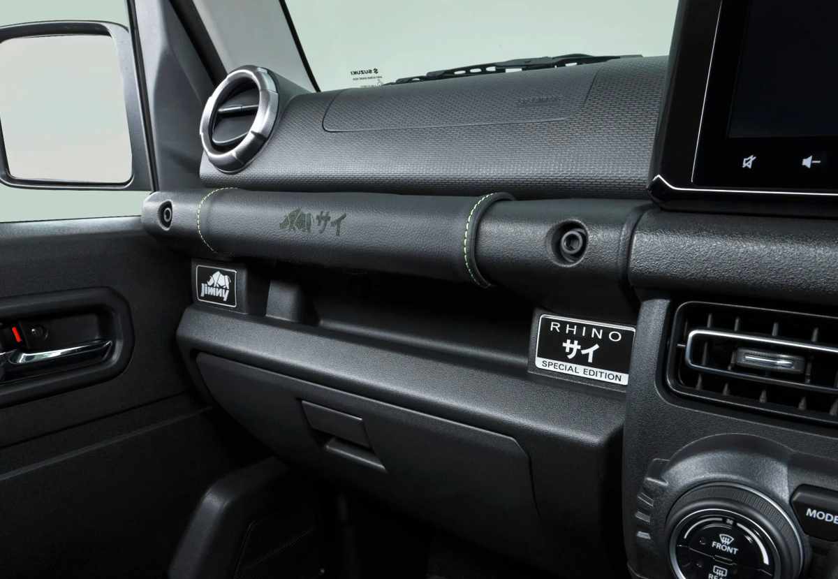 Primer plano de la agarradera frontal sobre el tablero del copiloto del Suzuki Jimny Rhino, forrada en cuero negro con costuras amarillas y la palabra Rhino grabada con láser.