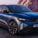 Vista frontal de tres cuartos del nuevo Renault Mégane E-Tech 2027 color azul satinado estacionado junto a la costa, destacando su nueva parrilla oscura y luces diurnas verticales.