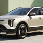 Vista frontal tres cuartos de la nueva SUV híbrida Kia Niro 2027 color beige estacionada frente a un edificio moderno a la luz del día, destacando sus nuevas luces diurnas LED en forma de T y rines aerodinámicos.