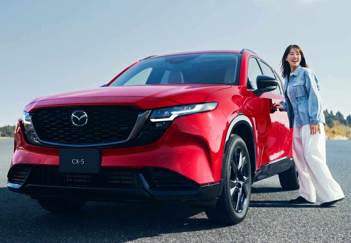 Vista frontal de la nueva Mazda CX-5 en color rojo brillante en una pista abierta, con una mujer sonriente vestida con chaqueta de jean abriendo la puerta del conductor del vehículo.