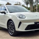 Vista frontal tres cuartos de la nueva SUV eléctrica GWM Ora 5 en color blanco marfil, estacionada sobre un camino de piedra rodeado de jardines, destacando su capó curvo y sus característicos faros redondos de estilo retro.