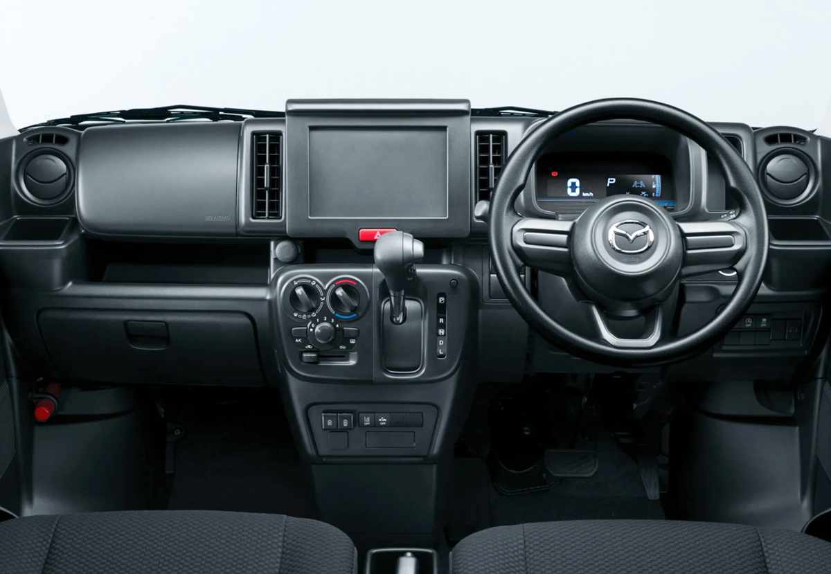 Tablero de instrumentos interior de la furgoneta Mazda Scrum en color negro, exhibiendo el nuevo volante multifunción de tres radios, la ubicación central de la palanca de cambios y una gran cubierta plástica en el espacio de la radio.