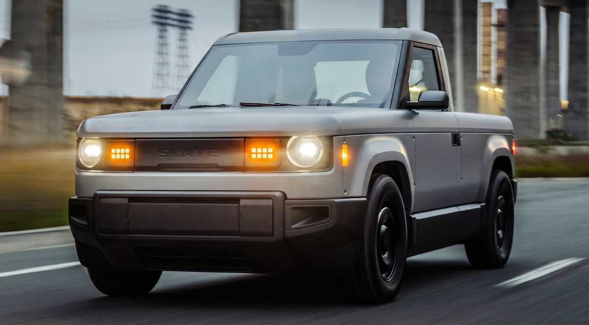 Camioneta eléctrica Slate Truck color gris rodando por autopista, mostrando su diseño frontal con faros redondos e iluminación LED naranja, destacando como el carro eléctrico barato americano.
