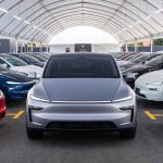 Vehículo eléctrico Tesla color plata en primer plano, rodeado por decenas de carros de la marca en distintos colores dentro de una gran carpa de exhibición.