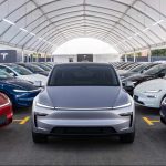Vehículo eléctrico Tesla color plata en primer plano, rodeado por decenas de carros de la marca en distintos colores dentro de una gran carpa de exhibición.