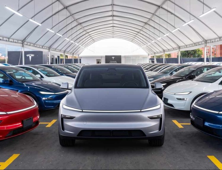 Vehículo eléctrico Tesla color plata en primer plano, rodeado por decenas de carros de la marca en distintos colores dentro de una gran carpa de exhibición.