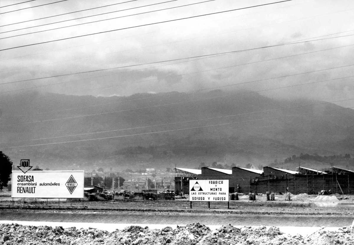Fotografía antigua en blanco y negro del paisaje montañoso y el letrero publicitario durante la construcción original de la planta de Sofasa en Envigado, Antioquia.