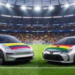 Un Tesla Model Y plateado con la bandera de Colombia en el capó y un Toyota Corolla gris con la bandera de Ghana se enfrentan en la cancha de un estadio de fútbol lleno de hinchas durante el Mundial 2026.
