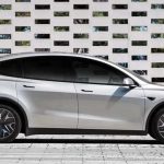 Vista lateral de perfil completo de la nueva Tesla Model Y L color plata en su variante extendida, estacionada frente a una pared geométrica blanca.