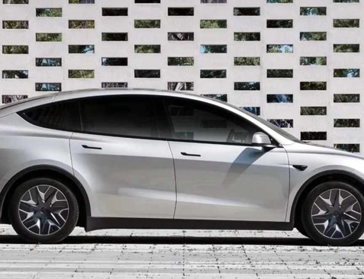 Vista lateral de perfil completo de la nueva Tesla Model Y L color plata en su variante extendida, estacionada frente a una pared geométrica blanca.