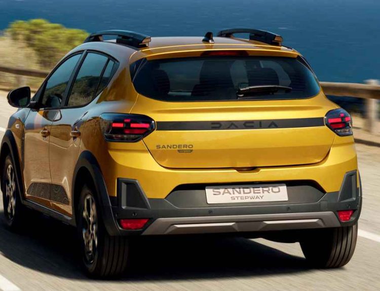 Vista trasera en movimiento del Dacia Sandero Stepway híbrido color amarillo, circulando por una carretera costera iluminada por el sol, destacando sus barras de techo y luces LED.