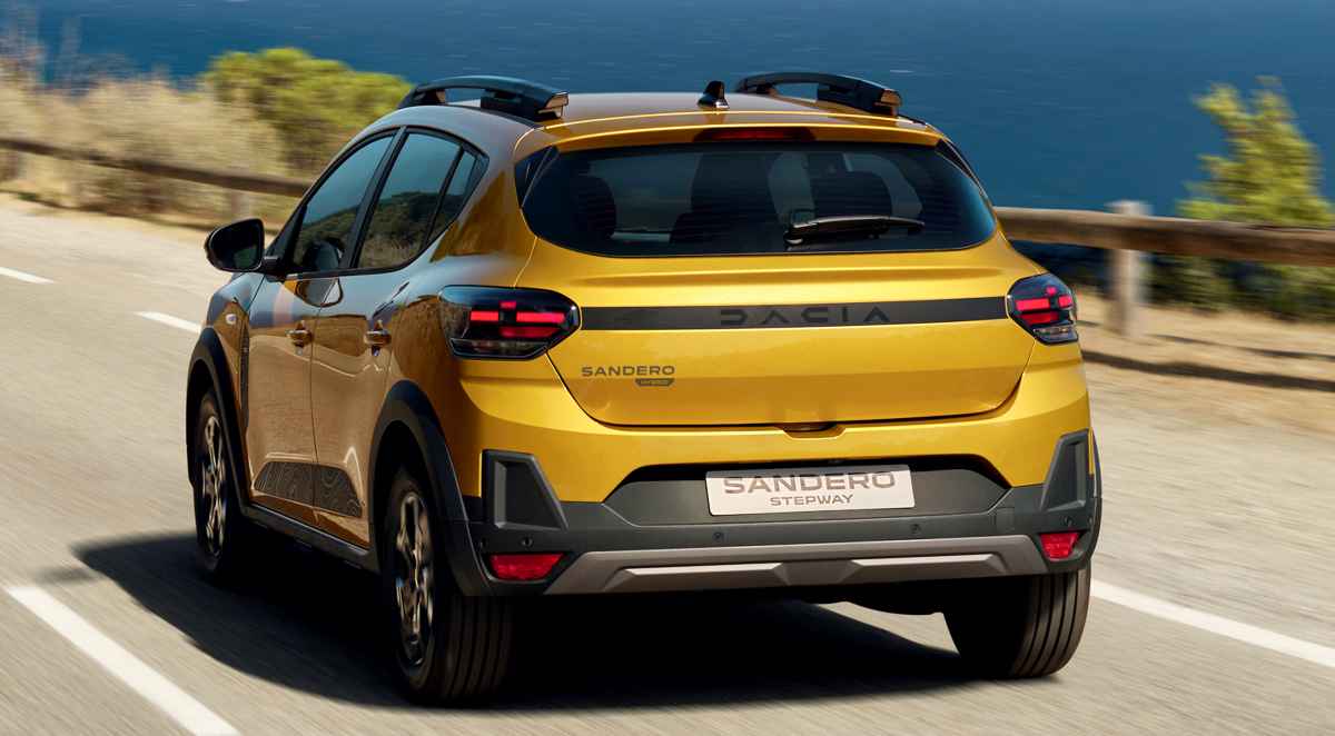 Vista trasera en movimiento del Dacia Sandero Stepway híbrido color amarillo, circulando por una carretera costera iluminada por el sol, destacando sus barras de techo y luces LED.