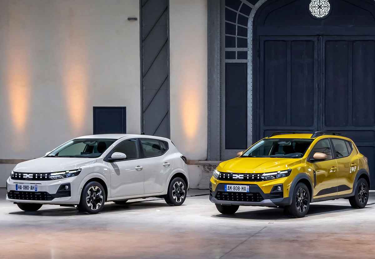 Vista frontal de tres cuartos de los nuevos Dacia Sandero blanco y Sandero Stepway amarillo con tecnología híbrida, exhibidos juntos bajo iluminación cálida en un entorno industrial.