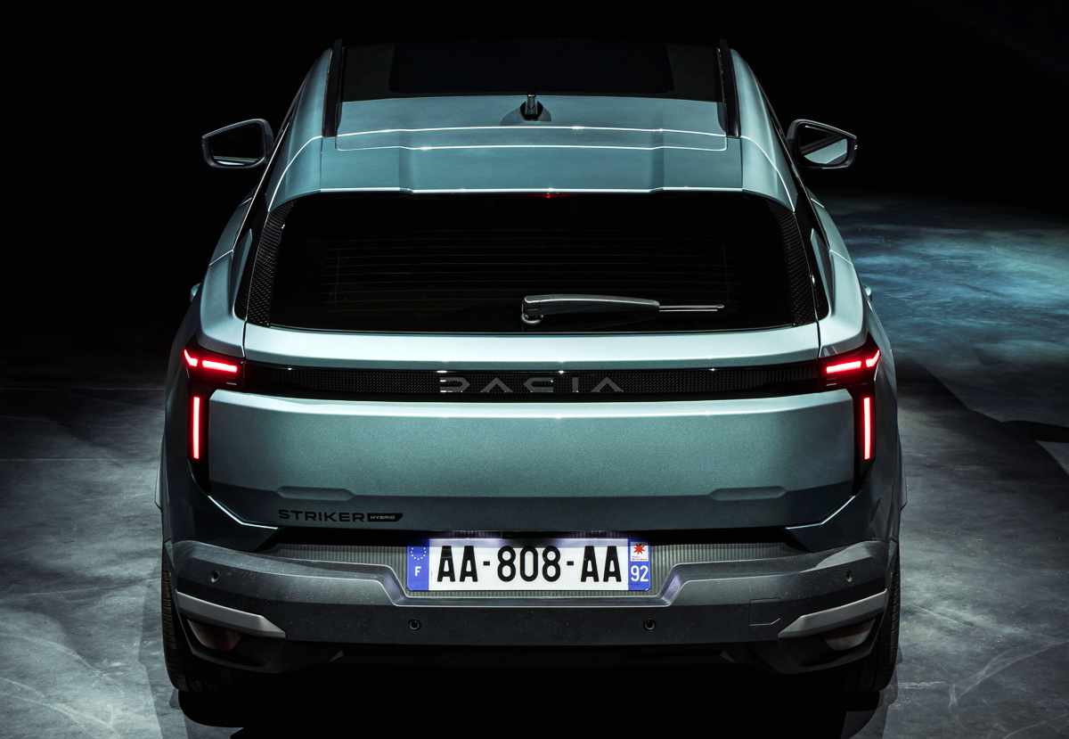 Toma trasera directa y simétrica de la camioneta SUV Dacia Striker en un tono verde-plateado oscuro metálico, con las luces traseras LED continuas encendidas. La franja negra central de las luces muestra la marca 'DACIA' en el centro. Debajo de la luz trasera izquierda, hay una insignia que dice 'STRIKER' y debajo de esta 'HYBRID'. La placa de matrícula francesa tiene el texto 'AA-808-AA'. También se aprecian las barras de techo plateadas sobre el techo y el limpiaparabrisas trasero.