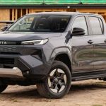 Vista frontal de tres cuartos de una Toyota Hilux color gris oscuro estacionada frente a una obra de construcción en madera, ilustrando su enfoque como pick-up para el trabajo duro.
