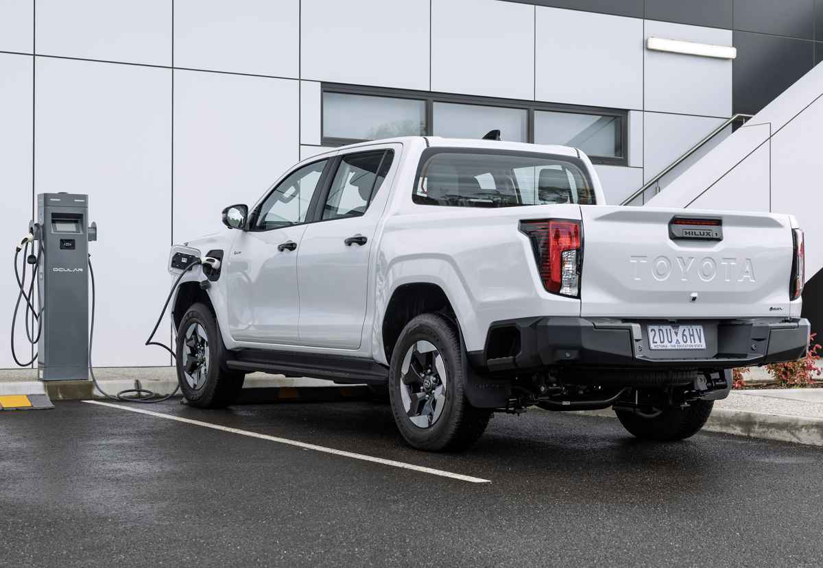 Vista trasera de tres cuartos de una pick-up Toyota color blanco conectada a una estación de carga eléctrica, representando la tecnología híbrida enchufable PHEV.
