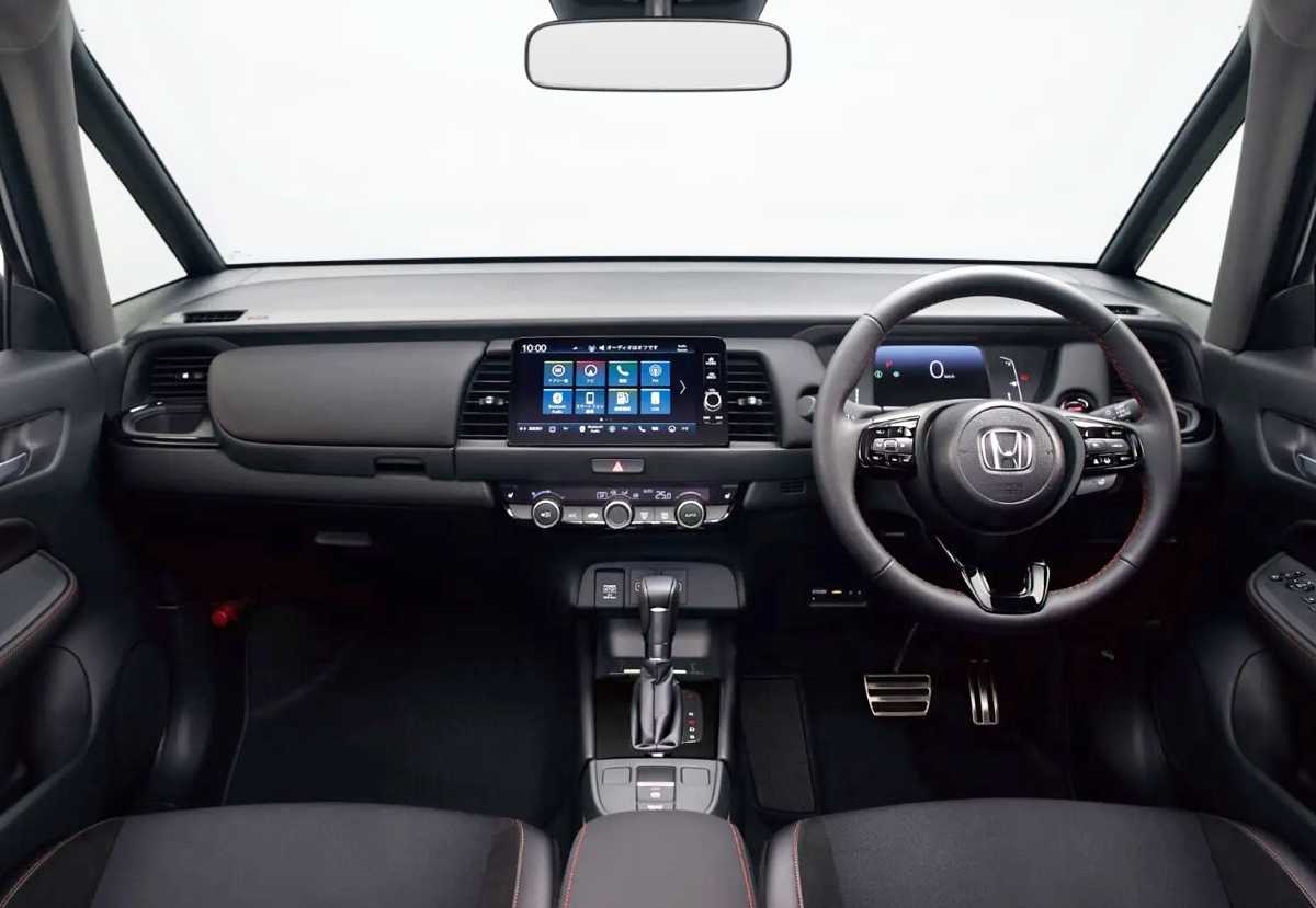 Habitáculo interior del renovado Honda Fit con volante a la derecha, mostrando el cuadro de instrumentos digital, la pantalla central de 9 pulgadas y sutiles costuras rojas.
