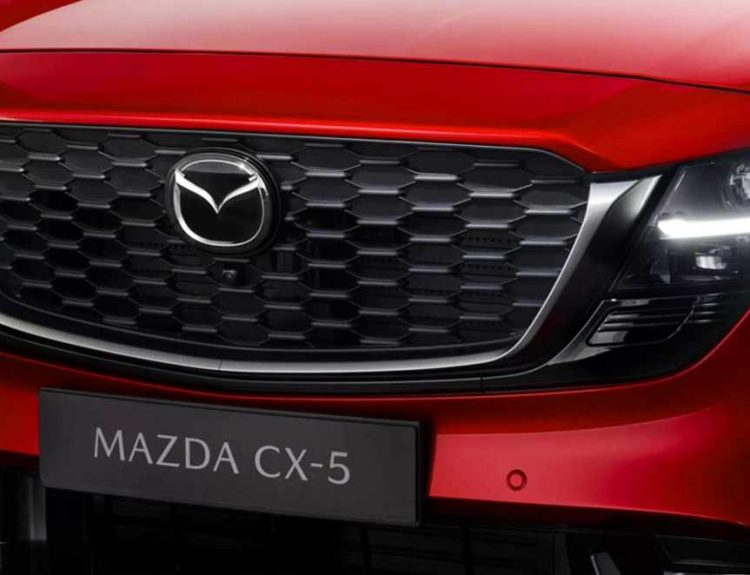 Detalle frontal cercano de la parrilla negra y el faro LED de la nueva Mazda CX-5 color rojo brillante, modelo que estrenará el primer sistema híbrido completo de la marca.