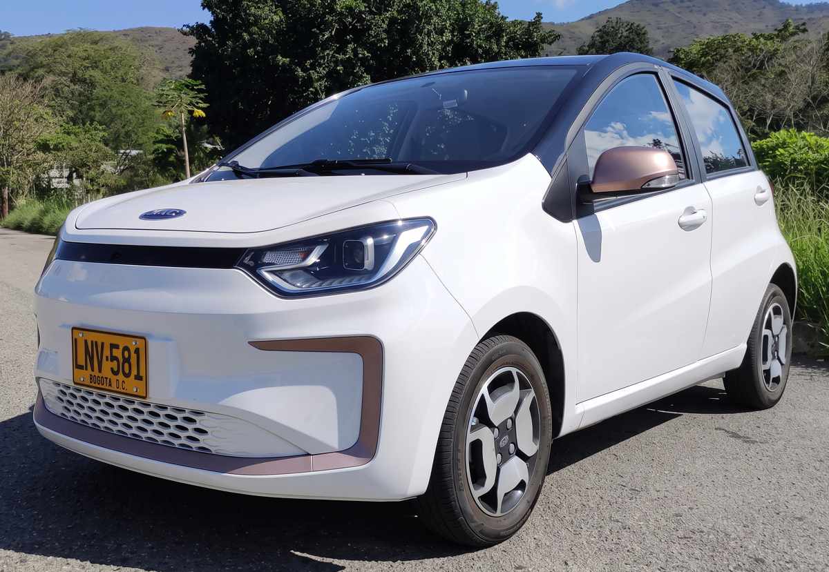 Vista frontal de tres cuartos del JAC E10X color blanco, un carro eléctrico urbano asequible, rodando por una vía pavimentada en las afueras de la ciudad.