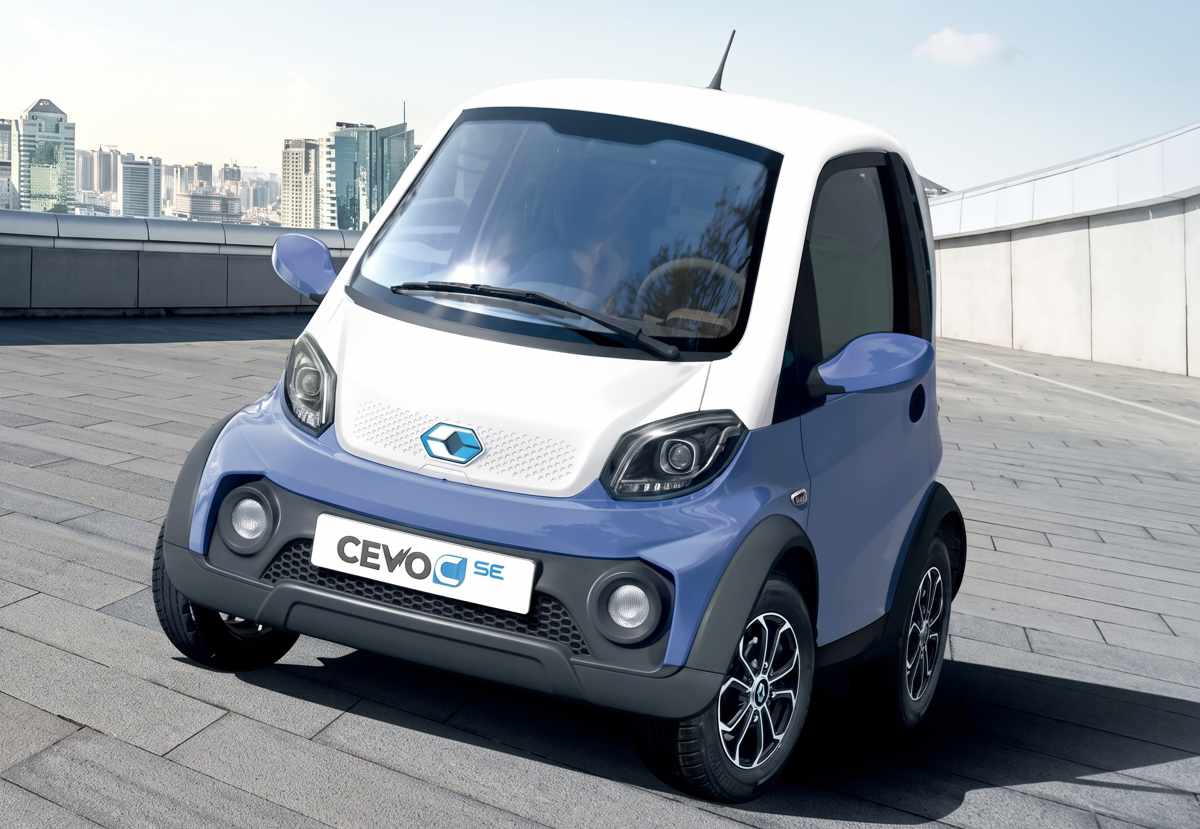 Cuadriciclo eléctrico CEVO C SE con carrocería bitono azul y blanco, diseñado para trayectos cortos, estacionado en una plaza con arquitectura moderna.