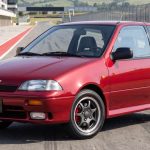 Vista frontal de tres cuartos del clásico Chevrolet Swift GTi color rojo estacionado en una pista de carreras, destacando su diseño deportivo, exploradoras y rines originales.