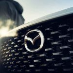 Detalle de la distintiva parrilla frontal y el emblema de Mazda iluminado por el sol, anticipando la llegada de su nueva línea de vehículos híbridos.