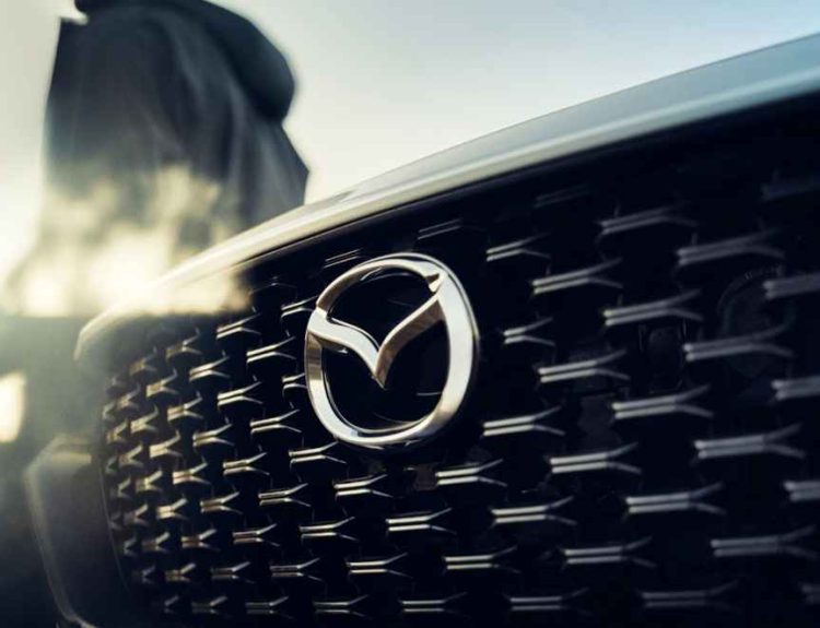 Detalle de la distintiva parrilla frontal y el emblema de Mazda iluminado por el sol, anticipando la llegada de su nueva línea de vehículos híbridos.