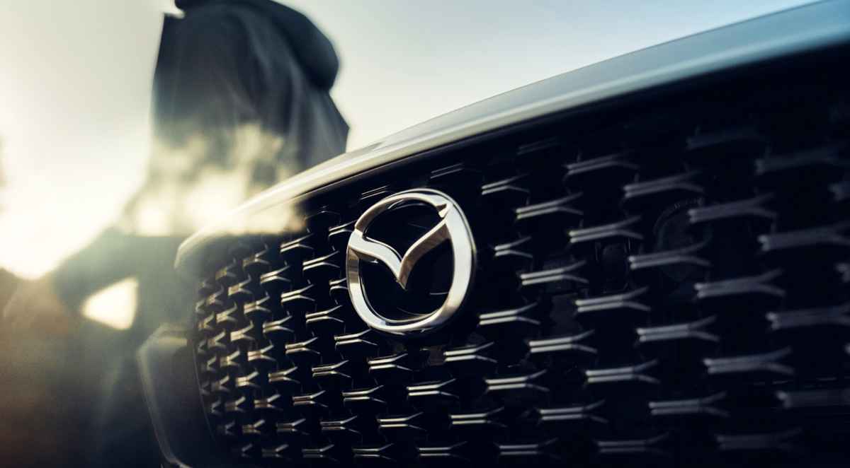 Detalle de la distintiva parrilla frontal y el emblema de Mazda iluminado por el sol, anticipando la llegada de su nueva línea de vehículos híbridos.