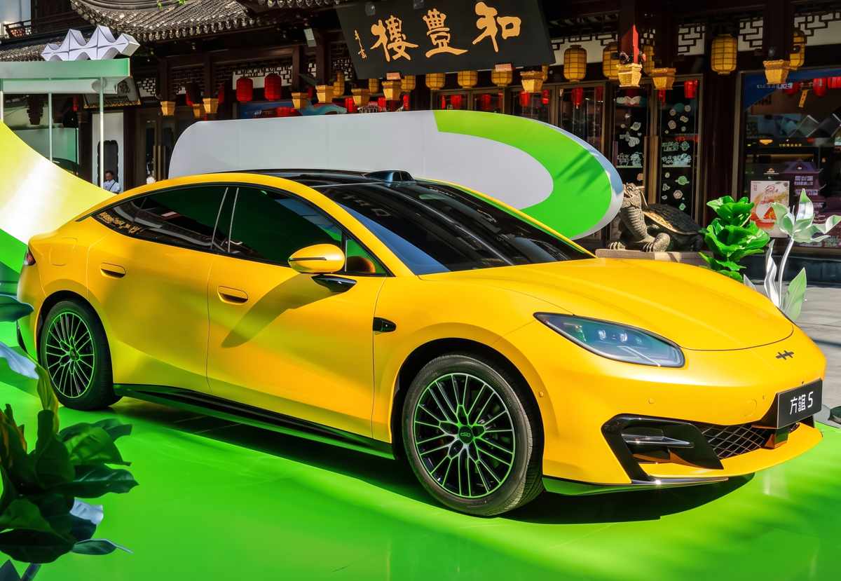 Exhibición del sedán eléctrico BYD Fang Cheng Bao Formula S color amarillo, estacionado sobre una plataforma verde brillante en un entorno urbano con arquitectura tradicional china.