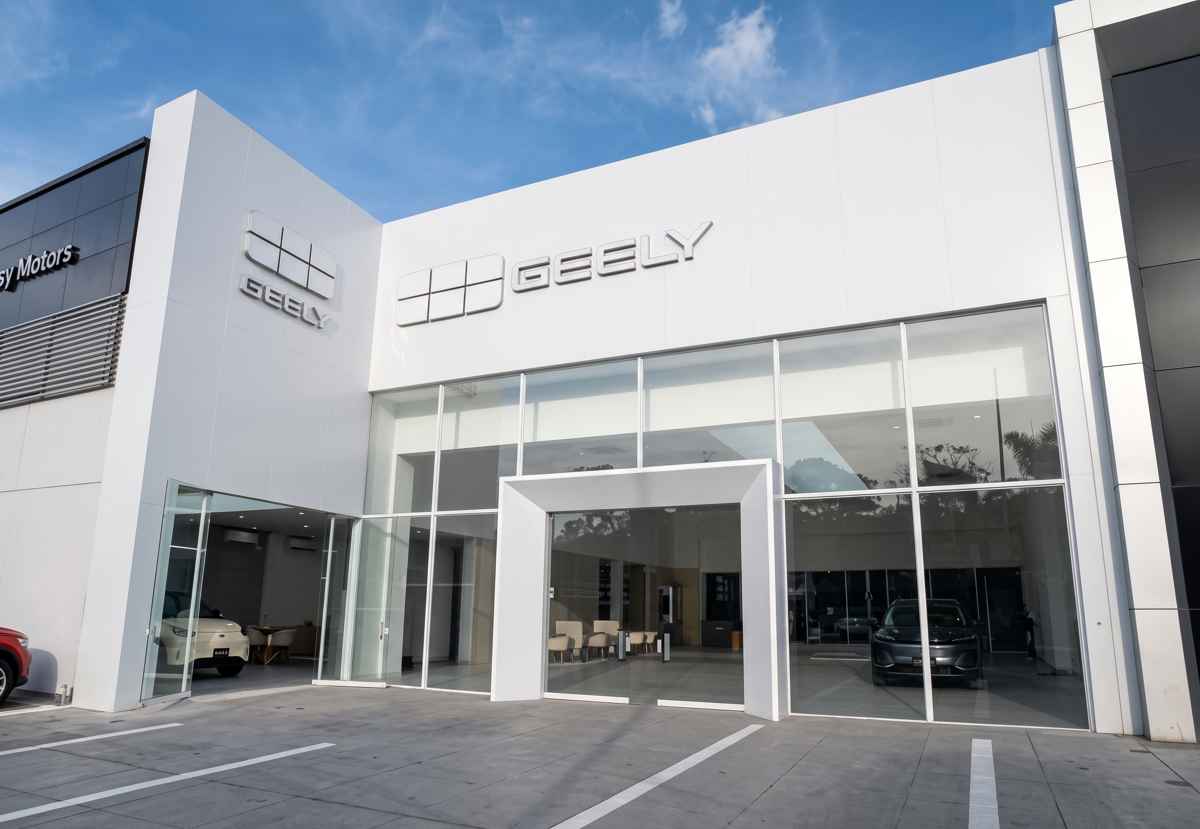 Fachada exterior de color blanco del nuevo concesionario Geely en alianza con Massy Motors, mostrando su entrada principal de cristal y vehículos de exhibición en el interior.