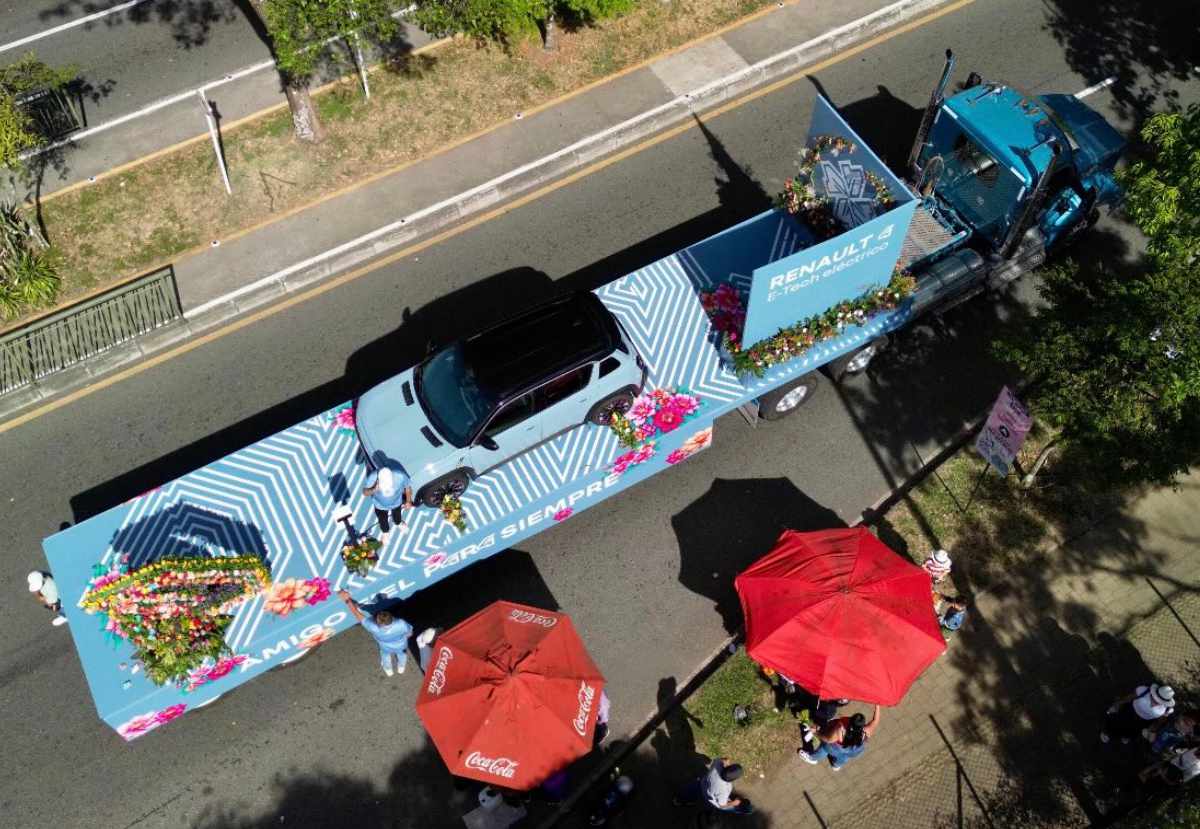 Fotografía aérea de un camión azul remolcando la plataforma con el nuevo Renault 4 eléctrico, desfilando por una amplia avenida rodeada de público y sombrillas rojas.