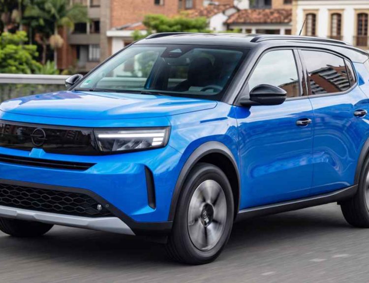 Vista frontal de tres cuartos de la nueva SUV Opel Crossland Híbrida color azul, rodando en movimiento por una avenida urbana con arquitectura clásica de fondo.