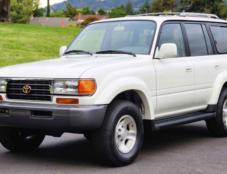 Vista frontal de tres cuartos de una impecable Toyota Land Cruiser Serie 80 'Burbuja' modelo 1997 color blanco, exhibida sobre asfalto, destacando su estado original y la parrilla frontal con el emblema dorado.