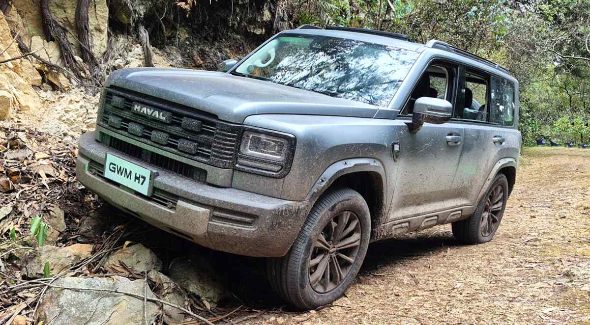 Plano frontal de tres cuartos de la SUV GWM Haval H7 Full Hybrid color gris oscuro superando un obstáculo rocoso en un camino de tierra, destacando su robusta parrilla y capacidades 4WD.