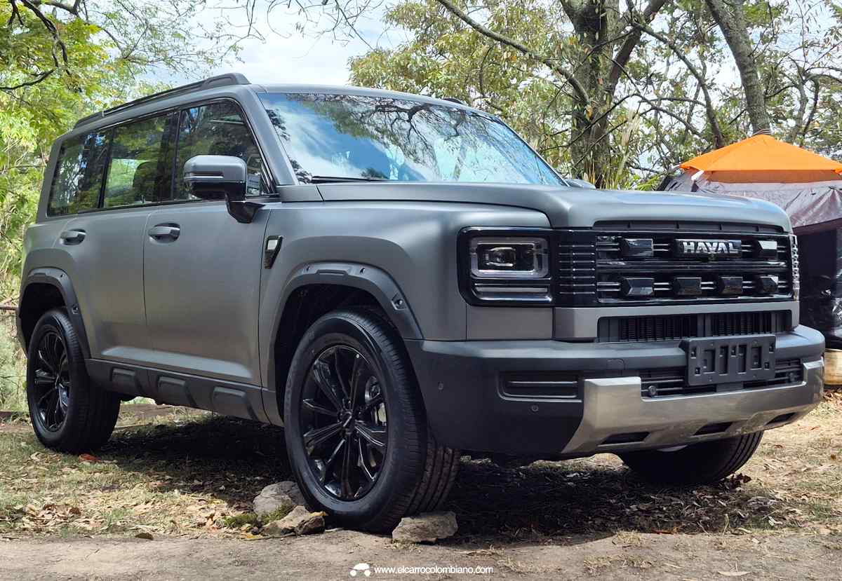 Plano frontal de tres cuartos de la GWM Haval H7 Full Hybrid color gris mate estacionada en una zona boscosa de camping, exhibiendo sus faros cuadrados retro y pasos de rueda anchos.