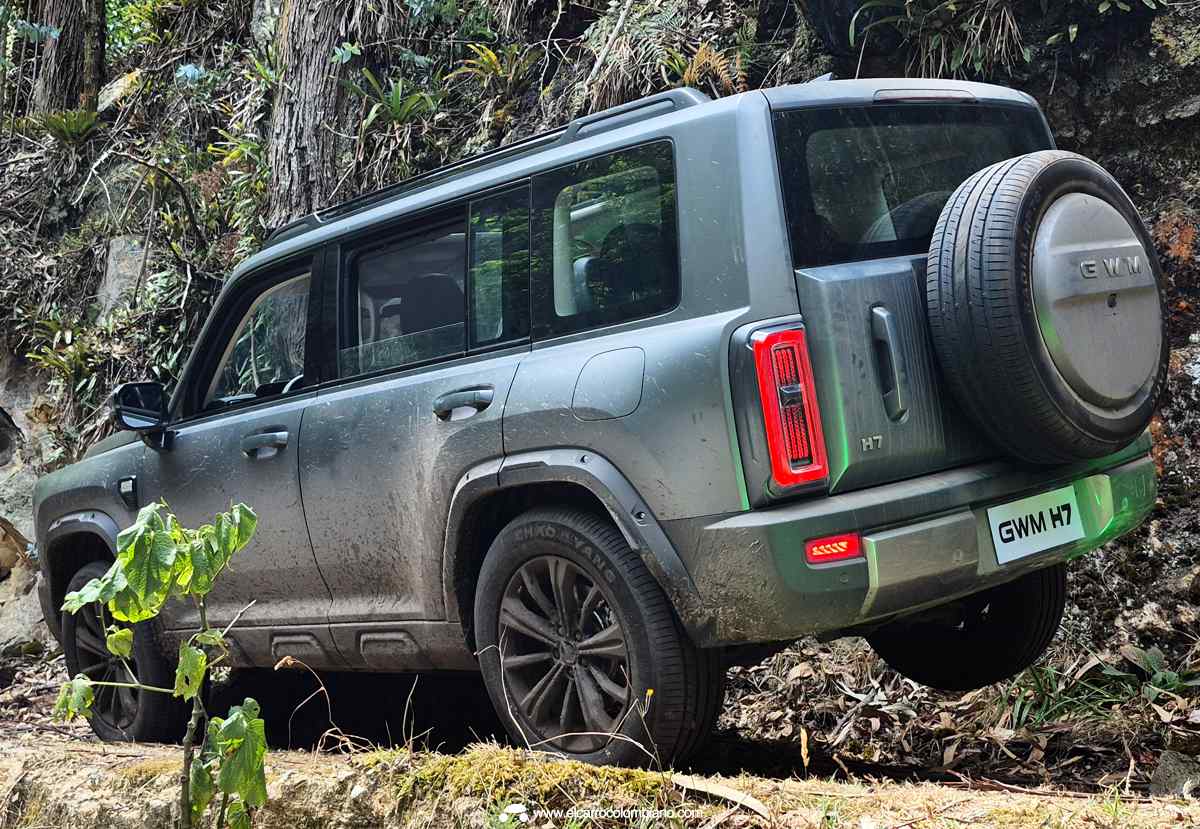 Plano de tres cuartos trasero de la GWM Haval H7 Full Hybrid color gris mate cubierta de barro sobre un terreno pedregoso, mostrando su portón de apertura lateral con llanta de repuesto.