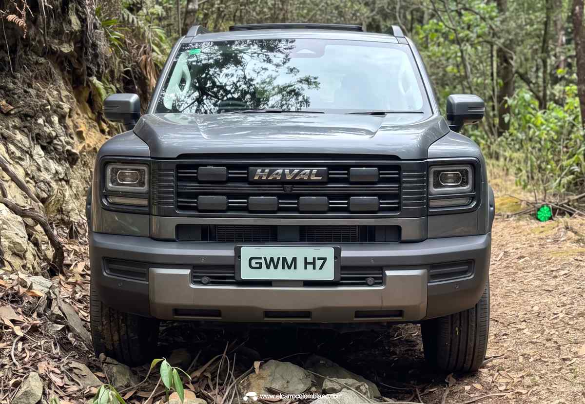 Plano frontal directo de la GWM Haval H7 Full Hybrid color gris mate sobre un camino boscoso, destacando la inscripción HAVAL en la parrilla rectangular y las prominentes luces exploradoras.