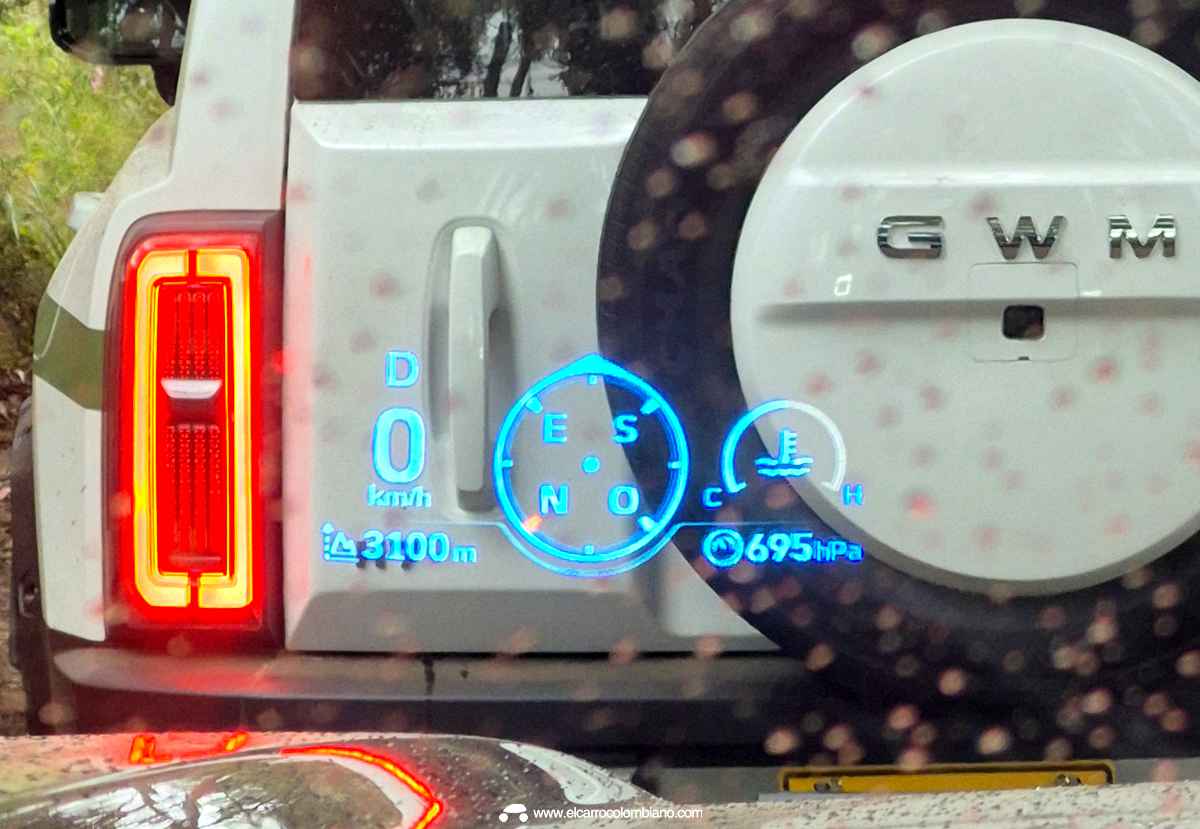 Plano detalle del Head-Up Display (HUD) proyectado en el parabrisas de la GWM Haval H7 Full Hybrid bajo la lluvia, mostrando los indicadores digitales de velocidad, altitud y presión, con otra H7 blanca visible al fondo.