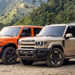 Plano general de las nuevas SUV iCAUR V23 color naranja e iCAUR V27 color arena estacionadas juntas sobre un camino de tierra, con palmas de cera y montañas verdes al fondo.