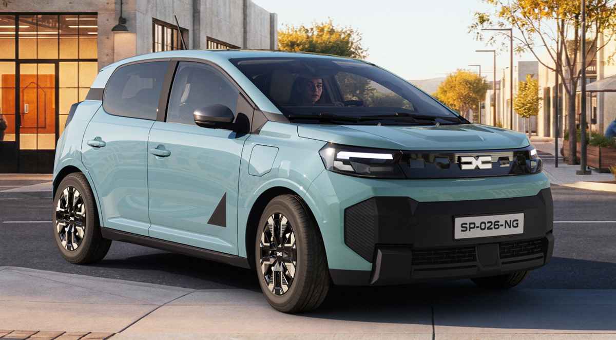 Plano frontal de tres cuartos del Dacia Spring color azul claro circulando por una calle urbana iluminada, destacando su renovada parrilla negra con el nuevo emblema de la marca.