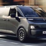 Plano frontal de tres cuartos cercano de la Renault Trafic E-Tech eléctrica gris en movimiento sobre una vía urbana, destacando su capó corto, firma luminosa LED con el rombo integrado y carrocería de carga.